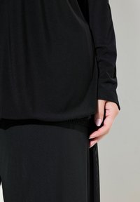 Schwarzes Oberteil mit langen Ärmeln, aus einem weichen, strukturierten Stoff, in entspannter Passform, über einem passenden schwarzen Rock. Eine Hand ruht auf dem Rock.