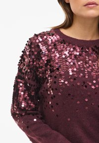 Maroon pulover okrašen z bleščicami, ki ima teksturirano pletenino, okrogel izrez in oprijeto silhueto s svetlečimi, okroglimi bleščicami na ramenih.