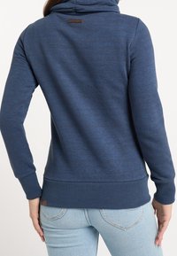 Námořnická modrá mikina s kapucí, má ribbované manžety a lem. Obsahuje texturovanou tkaninu a malou značkovou visačku na zadní straně blízko výstřihu.