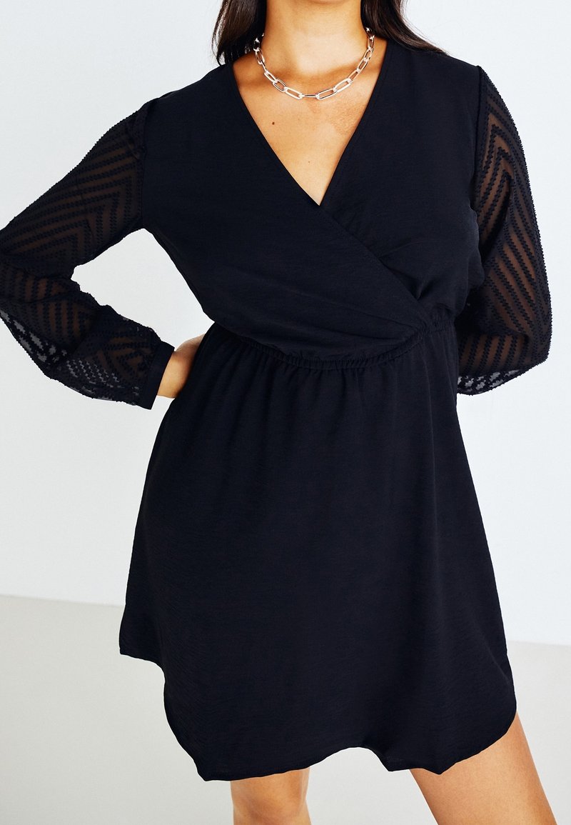 Femme portant une robe noire avec des manches longues transparentes à motifs et un décolleté en V, accessoirisée d’un collier chaîne en argent, main sur la hanche.