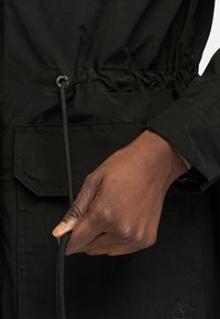 Czarna kurtka wykonana z gładkiego materiału, z elastycznym paskiem w talii, plecakiem ze sznurkiem oraz przednią kieszenią z klapą.