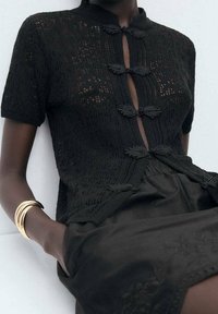 Schwarze gestrickte Strickjacke mit kurzen Ärmeln, ausgestattet mit einem hohen Kragen und dekorativen Verschlüssen. Kombiniert mit einem schwarzen Rock, der eine ausgefeilte Textur zeigt.