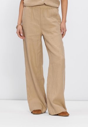 Vrouw draagt beige, loszittende linnen broek met elastische tailleband en bruine mocassins, staand tegen een effen achtergrond.