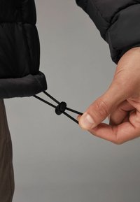 Main réglant un cordon élastique noir avec un verrou en plastique sur le poignet d'une veste matelassée noire sur un fond gris.