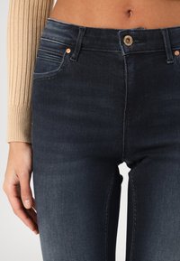 Dunkelblaue, hoch taillierte Jeans mit glatter Textur, dezenter Ausbleichung, goldfarbenen Beschlägen und Seitentaschendetails. Nahaufnahme der Vorderseite.
