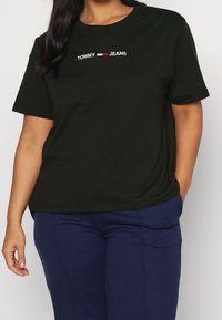 Camiseta de algodón negra con logo bordado "TOMMY JEANS" en blanco y rojo. Cuello redondo clásico, mangas cortas y corte relajado.