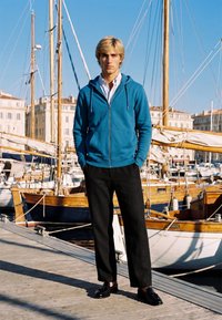 Jeune homme aux cheveux blonds debout sur un quai en bois, portant un sweat à capuche bleu, une chemise blanche et un pantalon noir, avec des voiliers et des bâtiments en arrière-plan.