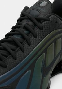 Gros plan sur une chaussure Nike Shox noire avec des lacets noirs et des panneaux latéraux en dégradé vert et bleu avec un logo Nike Swoosh noir.