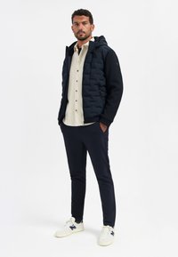 Marineblauwe gewatteerde jas met een textuurdesign, witte blouse en marineblauwe broek. Witte sneakers met blauwe accenten.