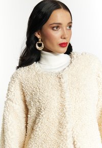 Îmbrăcăminte exterioară cu textură crem și finisaj pufoș, având un guler înalt confortabil dedesubt. Cerceii mari aurii completează look-ul.