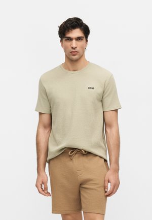 Férfi, aki bézs rövid ujjú inget visel "BOSS" logóval és világosbarna, zsinóros rövidnadrágot, egy egyszerű fehér háttér előtt áll.