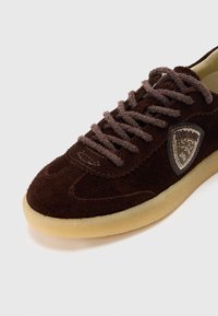 Σκούρο καφέ σουέντ αθλητικό παπούτσι με χοντρές κορδόνια στο ίδιο χρώμα, μπεζ λαστιχένια σόλα και στο πλάι διάκοσμος σε σχήμα ασπίδας με χάντρες.