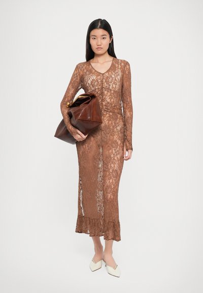 Robe en dentelle marron avec des manches longues, décolleté en V et ourlet volanté. Le modèle tient un grand sac en cuir marron avec un fermoir en or. Chaussures blanches à talons pointus.