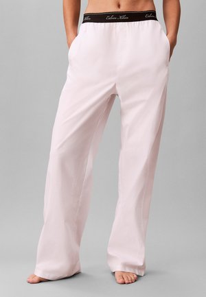 Persoon draagt lichtroze losse broek met zwarte Calvin Klein elastische tailleband, staat op blote voeten met handen in de zakken.