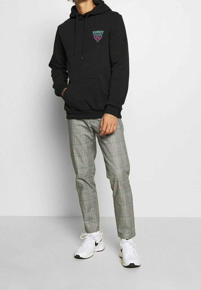 Personne portant un sweat à capuche noir avec le texte "Bored Inside", pantalon à carreaux gris et baskets blanches Nike, debout sur un fond blanc uni.