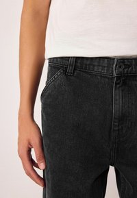 Schwarze Denim-Jeans mit einer Nahaufnahme einer vorderen Tasche, die eine subtile Textur und einen standardmäßigen Knopfverschluss zeigt.