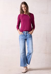 Magenta strukturierter Pullover mit langen Ärmeln, kombiniert mit hellblauen, weit geschnittenen Jeans. Das Model trägt hellbeige Slipper.