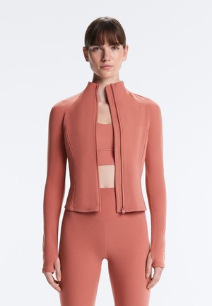 Žena nosi priliehavú, dlhorukávovú bundu na zips v farbe prachovej ruže cez zodpovedajúcu športovú podprsenku a legíny, stojaca na jednoduchom svetlom pozadí.