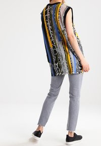 Haut sans manches multicolore avec des rayures verticales audacieuses en bleu, jaune, vert et noir, associé à un jean gris clair ajusté et des baskets noires.