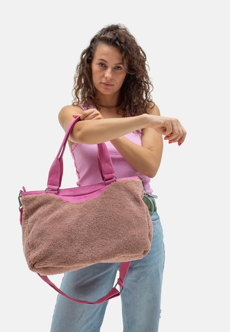 Femme aux cheveux bouclés portant un débardeur rose et un jean bleu clair, tenant un grand sac à épaule texturé rose et beige.