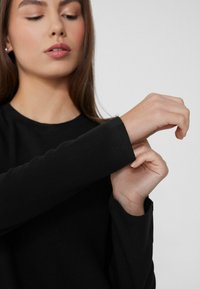Czarna bluzka z długim rękawem, z okrągłym dekoltem i dopasowanymi rękawami. Gładka tekstura o prostym wzorze, podkreślająca okolice nadgarstków.