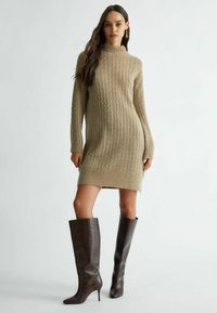 Abito maglione in beige chiaro con texture a coste, maniche lunghe e colletto alto. Abbinato a stivali alti fino al ginocchio in marrone scuro con motivo coccodrillo.