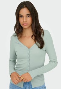 Jasnozielony kardigan z długim rękawem o prążkowanej fakturze, z dekoltem w serek i zapięciem na guziki. Posiada falbankowaty dół i dopasowany krój.
