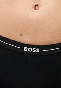 Κάτω κοιλιακή χώρα με ελιά πάνω από τη μαύρη ζώνη εσωρούχου που φέρει το «BOSS» σε λευκά γράμματα και δύο λεπτές λευκές οριζόντιες γραμμές.