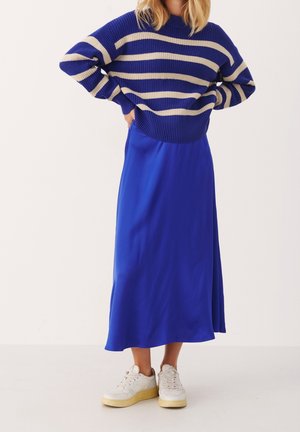 Pull en tricot bleu rayé avec des rayures beiges, associé à une jupe en satin bleu uni et des baskets blanches à semelles jaunes.