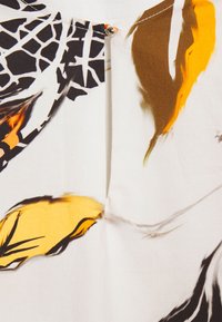 Tissu blanc avec des motifs abstraits noirs, jaunes et orange, avec un petit bouton-pression en métal près de la couture centrale supérieure.