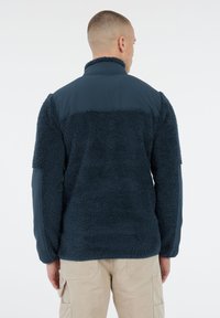 Man in een marineblauwe fleecejas met verstevigde stofpatches op de schouders en ellebogen, gecombineerd met beige cargo-broeken, staand met de rug naar de camera gekeerd.