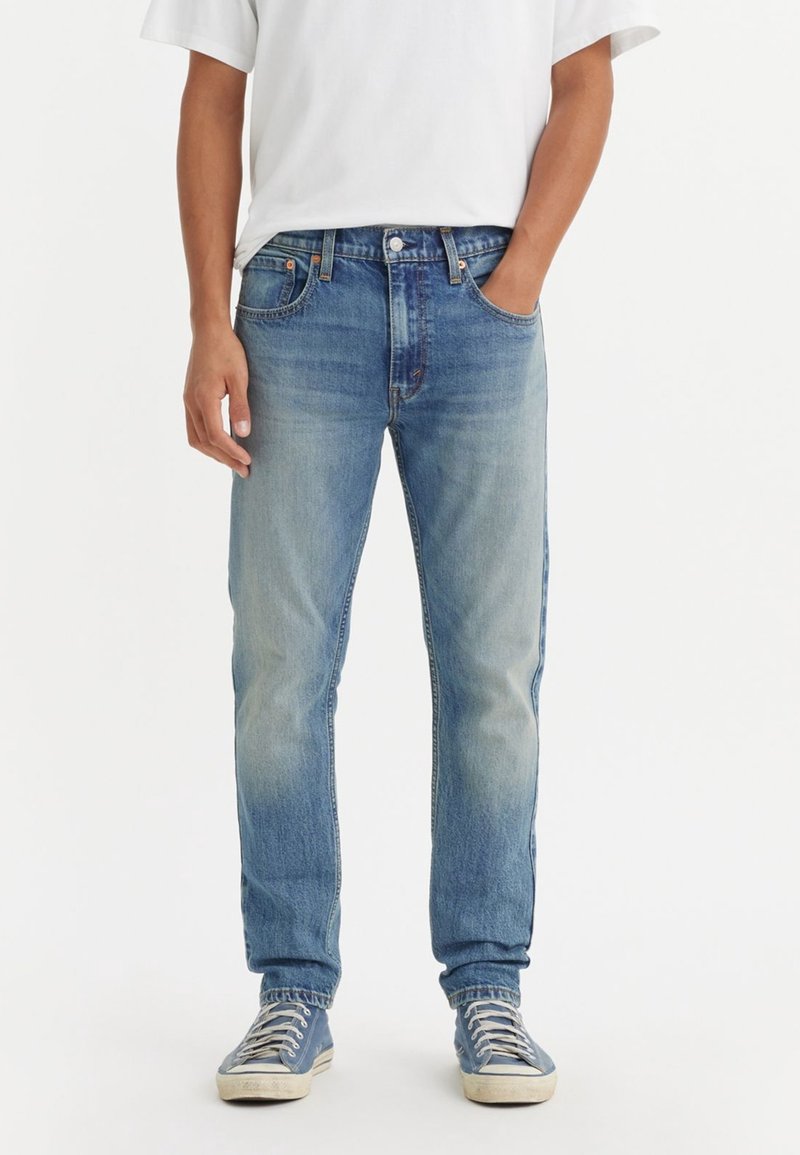 Homme portant un jean bleu clair délavé et des baskets bleues avec des lacets blancs, debout avec une main dans la poche et un t-shirt blanc.