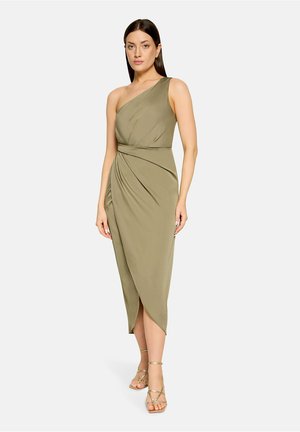 Kobieta ubrana w oliwkową, bezrękawną sukienkę midi z jednym odkrytym ramieniem i marszczeniami, w połączeniu ze złotymi sandałami na paski.