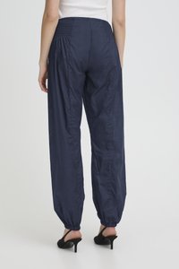 Moteris dėvi tamsiai mėlynas elastingas kelnaites su rankogaliais, derinamas su juodais aukštakulniais su atvira kulno dalimi, stovinti priešais paprastą šviesią foną.