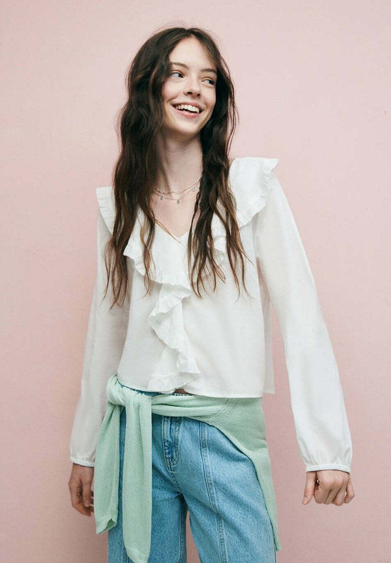 Jeune femme souriante, portant un chemisier blanc à volants, un jean bleu clair et un pull vert clair noué autour de la taille, sur un fond rose.