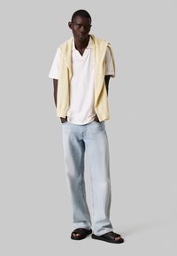 Witte polo onder een lichtgele trui, gecombineerd met lichtblauwe jeans en zwarte sandalen, staand tegen een grijze achtergrond.