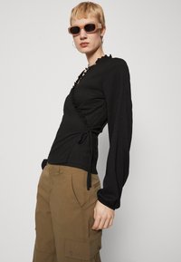 Bluză neagră înfășurată, cu detalii cu volane, mâneci lungi, dintr-un material texturat. Asortată cu pantaloni cargo khaki, cu buzunare laterale și o croială lejeră.