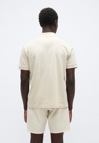 Μπεζ βαμβακερό πόλο πουκάμισο με κοντά μανίκια, σε άνετη εφαρμογή και με ελαφριά υφή, συνδυασμένο με ταιριαστά μπεζ σορτς. Πλάγια όψη από πίσω.