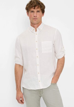 Weißes Leinenhemd mit Button-Down-Kragen, einer Brusttasche und hochgerollten Ärmeln. Kombiniert mit hellen Hosen.