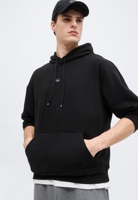 Noor mees musta kapuutsiga, käed ees taskus, must nokamüts peas, hallid püksid ja valge särk kapuutsi äärtest all paistmas.