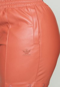 Terracotta leather shorts with elastic waistband, side pocket, and embossed Adidas logo. Features snap detail at the hem.