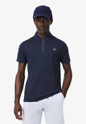 Uomo con un berretto blu navy e una polo con zip coordinata con un piccolo logo coccodrillo, abbinata a pantaloncini bianchi, in piedi con una mano in tasca.