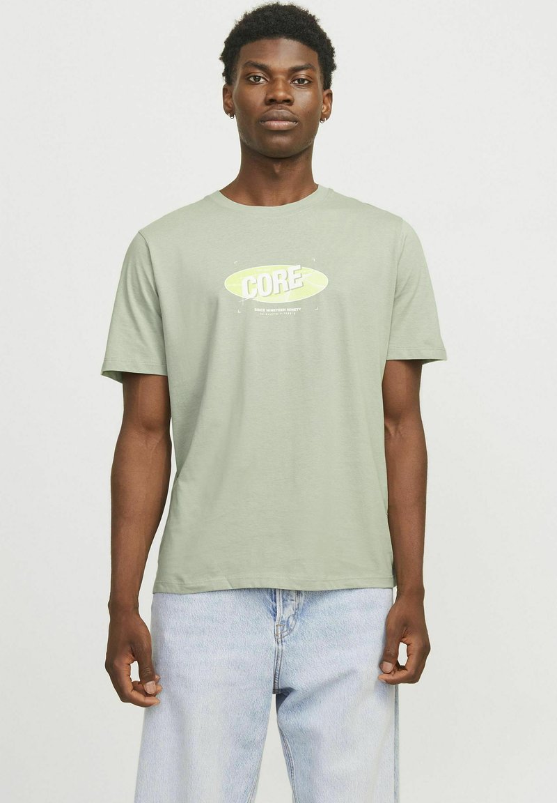 Camiseta de algodón verde claro con un gráfico central que dice "CORE" en amarillo y blanco; cuello redondo, mangas cortas y dobladillo recto. Combinada con jeans azul claro.