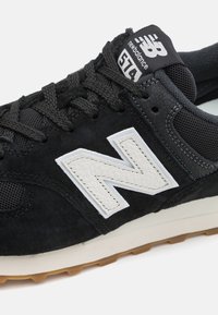 Black suede athletic shoe with mesh accents. Features a white leather "N" logo, textured midsole, and brown rubber outsole. Model 574.
