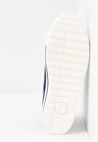 Semelle en caoutchouc blanche d'une chaussure marine montrant un motif de semelle, un détail circulaire près du talon, et la marque "Gabor", inclinée contre un fond blanc.