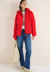 Rote Pufferjacke mit abnehmbarer Kapuze, strukturiertem Stoff, kombiniert mit hellblauen Jeans und einer cremefarbenen Tasche. Legere Schuhe runden den Look ab.