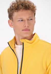 Rumena zip-up bunda z visokim ovratnikom, s pomirjujočim mornarsko modrim šivom. Nosi se čez belo srajco, ki prikazuje gladko in mehko teksturo.