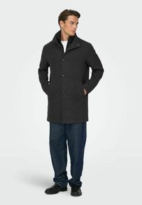 Szary płaszcz z mieszanki wełny z wysokim kołnierzem, guzikami z przodu i bocznymi kieszeniami, zestawiony z ciemnymi spodniami dżinsowymi i czarnymi butami.
