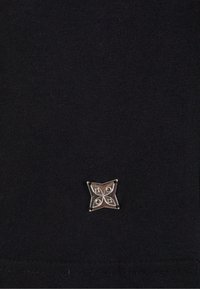 Melna adīta auduma ar mazu, sudraba četru ziedlapu emblemu, kas satur centrālās detaļas. Audums ir mīksts un ar gludu tekstūru.