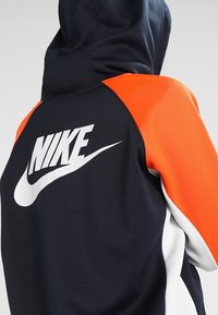 Felpa con zip blu navy e arancione, caratterizzata da un pannello arancione testurizzato sulla spalla, logo Nike bianco sul retro e cappuccio.